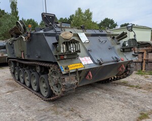 Military armored vehicle parked at a storage facility in a wooded area during daytime, showcasing its rugged design and camouflage features