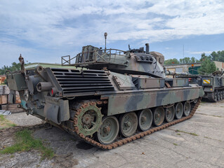 Military tank parked at a military facility surrounded by vehicles and equipment during daylight hours in a cloudy atmosphere