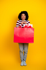 Cheerful young woman holding a vibrant red shopping bag while standing against a bright yellow...