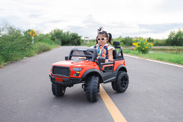 Fototapeta premium A cute little Asian girl is happily riding a small toy car along a country road,A girl drives a toy car on a concrete road.