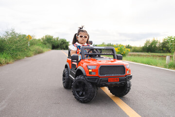 Fototapeta premium A cute little Asian girl is happily riding a small toy car along a country road,A girl drives a toy car on a concrete road.