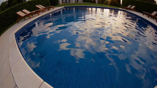 wide-angled panoramic shot of clean, reflective swimming pool surrounded by wooden loungers and lush gardens at high-end villa during the time of day characterized by warm, soft sunlight