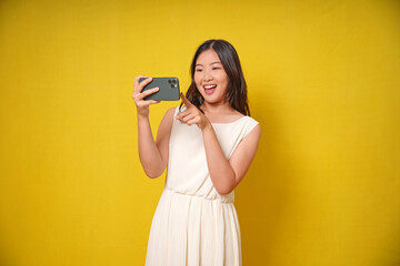 A cheerful Asian woman holding her smartphone and wearing a white dress poses cheerfully and smiles at the camera against an isolated yellow background.