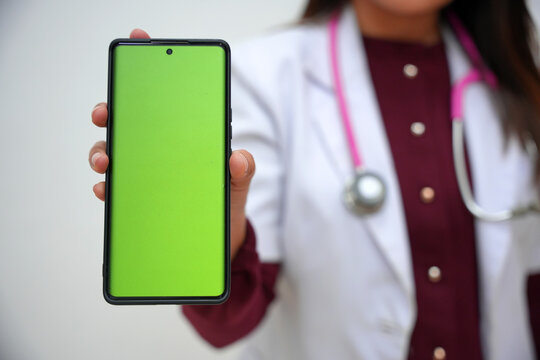 Charming Asian female doctor in a white gown shows a green screen mockup of a smartphone to add your graphic banner, standing against a white background. Recommend new apps, attractive promotions. - Powered by Adobe