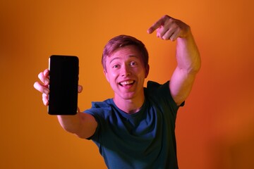 Happy man pointing at smartphone in neon lights against orange background