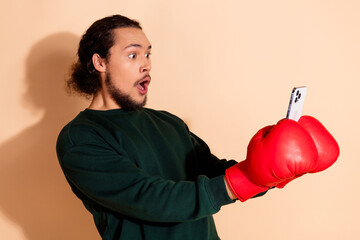 Man with boxing gloves holding a smartphone and expressing surprise against a beige background in a...