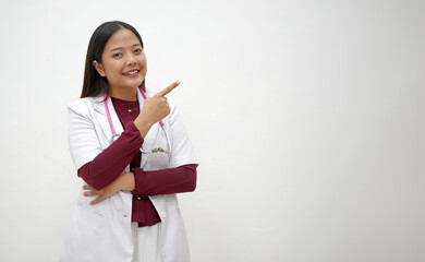 Cheerful Asian female doctor points up into empty space with one hand, showing excitement, wearing...