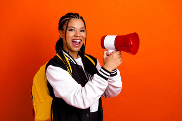 Cheerful teenage student holding megaphone, wearing a yellow backpack on orange background,...