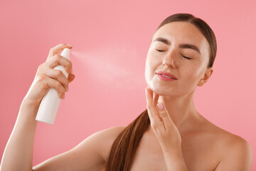 Beautiful woman spraying thermal water onto her face on pink background