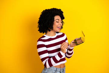 Young woman with curly hair holding a makeup palette on a vibrant yellow background, smiling while...
