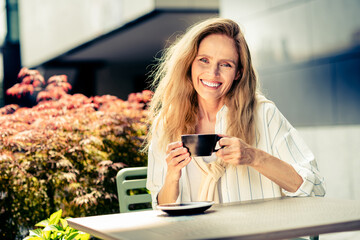 Middle-aged woman enjoying her morning coffee outdoors in a sunny urban setting filled with natural beauty