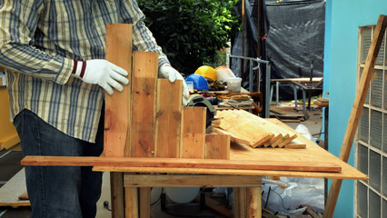 Carpenter wearing gloves arranging stacked wooden planks of different sizes on workbench in workshop