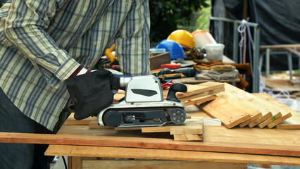 Worker sanding wooden plank with electric belt sander on workbench in outdoor carpentry workshop