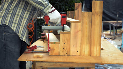 Worker in gloves using nail gun to fasten wooden planks into step-shaped structure during carpentry