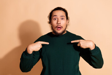 Young man with surprised facial expression pointing to himself wearing green sweatshirt posing...