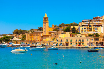 Colorful view of Marsaskala, Malta, with boats floating on the bright blue sea and historic stone buildings along the waterfront. A scenic coastal townscape under a clear summer sky