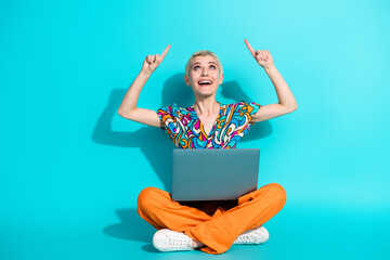 Excited young woman sitting cross-legged on teal background with laptop, pointing up cheerfully...