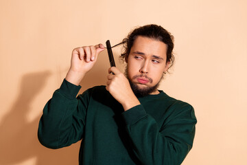Young man struggling with tangled hair while using a comb, beige backdrop, humorous moment of self-care, casual wear.