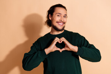 Cheerful man in casual green clothing forming a heart with hands on beige background, expressing...