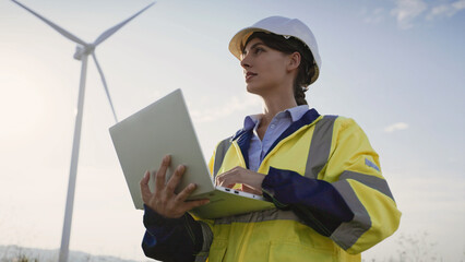 Cute Caucasian girl standing in front of working wind turbine. Entering information on laptop with one hand. Pressing keys on keyboard. Recording her observations as maintenance inspector.