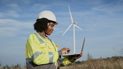 Focused African American woman looking around and recording data on laptop. Observing tall wind turbines. Inspector busy at work. Calculating possible ways of energy production improvement.