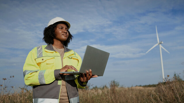 Side view of charming African American woman holding light laptop with one hand. Working comfortably with one hand in middle of open field. Power plant worker recording data. Sunny weather. - Powered by Adobe