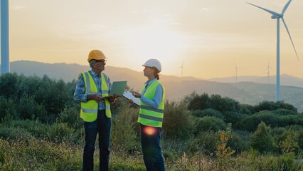 Two Caucasian coworkers standing before location with wind turbines. Engineers in working equipment discussing about something. Man using laptop while his business partner explaining his observations.