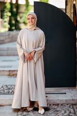 Naklejka premium Smiling woman in modest hijab and flowing dress adjusting scarf outdoors, standing near stone steps, symbolizing elegance, culture and modern lifestyle.