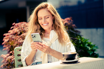 Charming mature woman enjoying leisure time outdoors with phone and coffee during a sunny day in the park