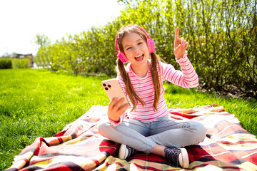 A cheerful young girl wearing headphones sits on a blanket outdoors, enjoying music and sunlight on...