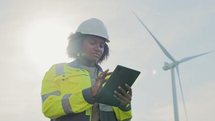Focused African American woman holding tablet device while tapping on screen with one finger. Recording her observations from checking wind turbine power plant. Protective equipment.