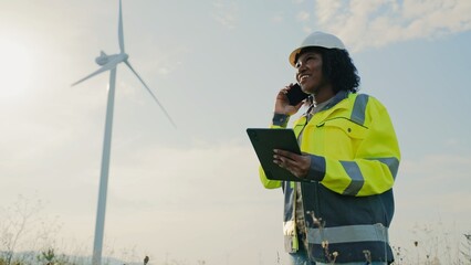 View from below of African American woman standing in middle of large field and talking on smartphone. Holding tablet device with one hand. Consulting with boss or manager. Wind turbine in background.
