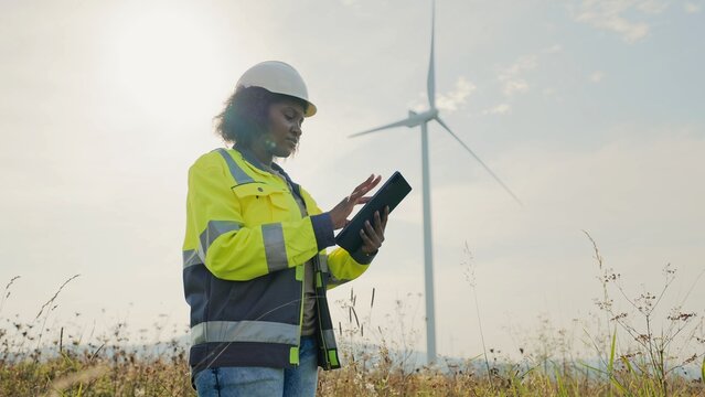 Charming African American woman completing inspection of working wind turbines. Checking quality of materials or production outcome. Recording data on tablet device. Blue sky in background.