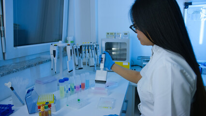 Asian female laboratory technician processes samples using a pipette surrounded by various lab...