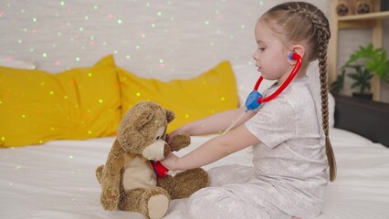 little child listens teddy bear with stethoscope, kid girl plays doctor while sitting on bed,...