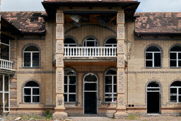 Abandoned brick mansion with arched windows, damaged roof, graffiti, and broken glass