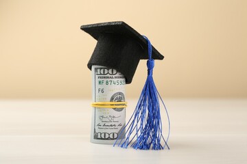 Scholarship. Graduate hat and dollar banknotes on white table against beige background, closeup