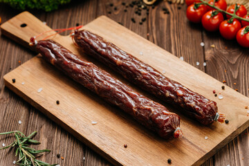 Dry cured sausage sticks on wooden board with herbs, tomatoes, and spices. Rustic food styling