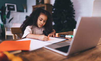 Fototapeta premium Focused cute ethnic girl writing in notepad while studying at table with laptop
