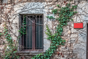 Rustic stone wall with barred window, ivy vines, and blank red sign. Weathered facade blends...