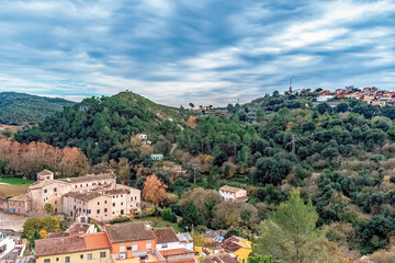 Obraz premium Scenic landscape of Girona, Spain with traditional stone buildings nestled in forested hills. Charming countryside view showing rural houses, green valleys, and a small village perched on the hilltop