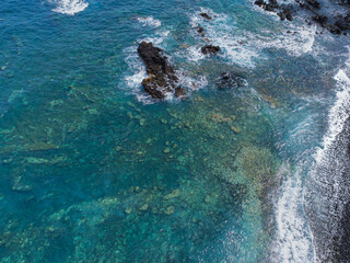 Aerial view of the rugged volcanic coast of Tenerife, Spain. Sharp, dark rock formations punctuate the vivid blue waters of the Atlantic Ocean. The jagged terrain creates natural pools 