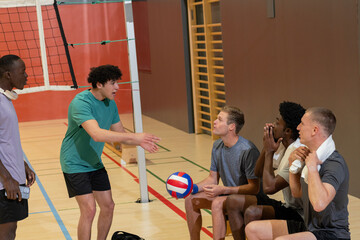 Diverse male teammates gesturing by net at gym holding ball smartphone headphones