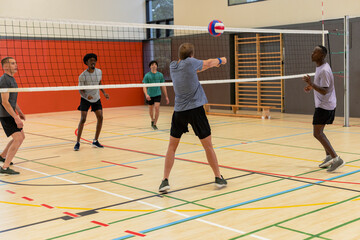 Diverse male teammates in sportswear playing volleyball in gym under net with ball