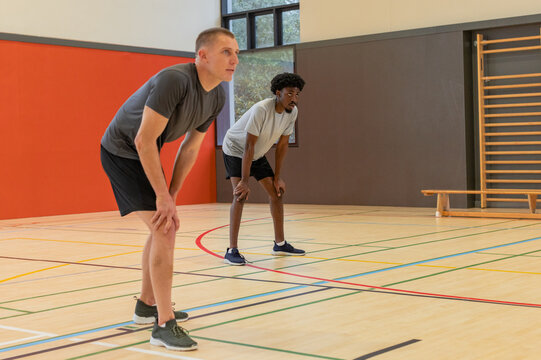 Diverse male friends bending forward and resting on court with colored lines inside gymnasium - Powered by Adobe