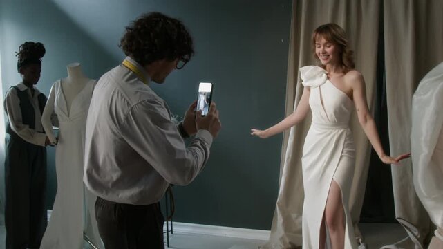 Bridal fashion designer taking photos of young woman in wedding dress on her phone during try-on