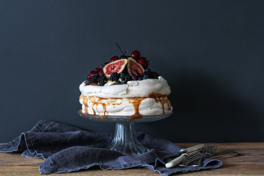 Pavlova dessert with figs, cherries, blueberries, blackberries, strawberries, whipped cream and toffee on a cakestand on a wooden table with cutlery
