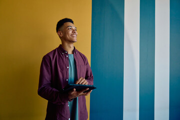Smiling young businessman looking away while using tablet against colorful wall in office