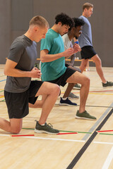 Diverse male workout teammates performing lunges in gymnasium in sportswear with dumbbells on bench