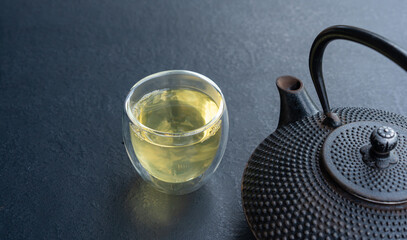 Glass cup with green tea and cast iron teapot on dark table, double wall glass